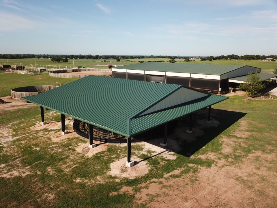 Aerial view of a green-roofed metal livestock shelter with open sides, located on a rural property with surrounding barns, fencing, and open fields.