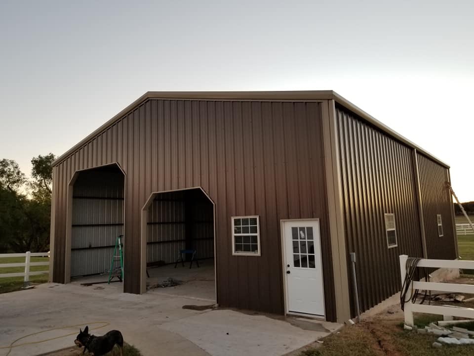 Partially completed brown metal building with two open garage bays, white entry door, and window, set on a concrete pad with fencing and a dog in the foreground.