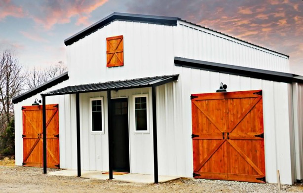 White metal barn-style building with black trim and striking wooden barn doors, set against a dramatic sunset sky.