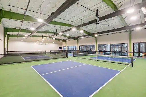 A professional photo shows the interior of a large, modern indoor sports facility. The space features three pickleball courts, two in the back and one in the foreground. The courts have blue playing surfaces and are marked with white lines, surrounded by a green floor. The walls are white, with a dark green border near the ceiling. Large industrial fans and bright lights hang from the high ceiling, and large windows line the back wall, letting in natural light. The name "Pickle House" is written on the back wall in gray text.