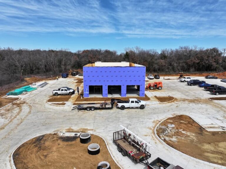 Construction site aerial view of a small commercial building wrapped in blue sheathing, surrounded by dirt and construction vehicles.