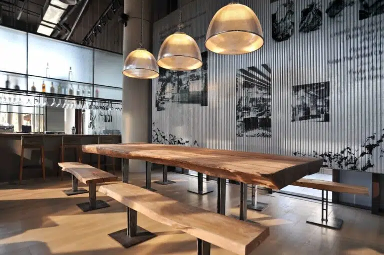 Modern restaurant or bar interior featuring a large live-edge wooden communal table, matching benches, industrial pendant lights, and corrugated metal wall paneling.